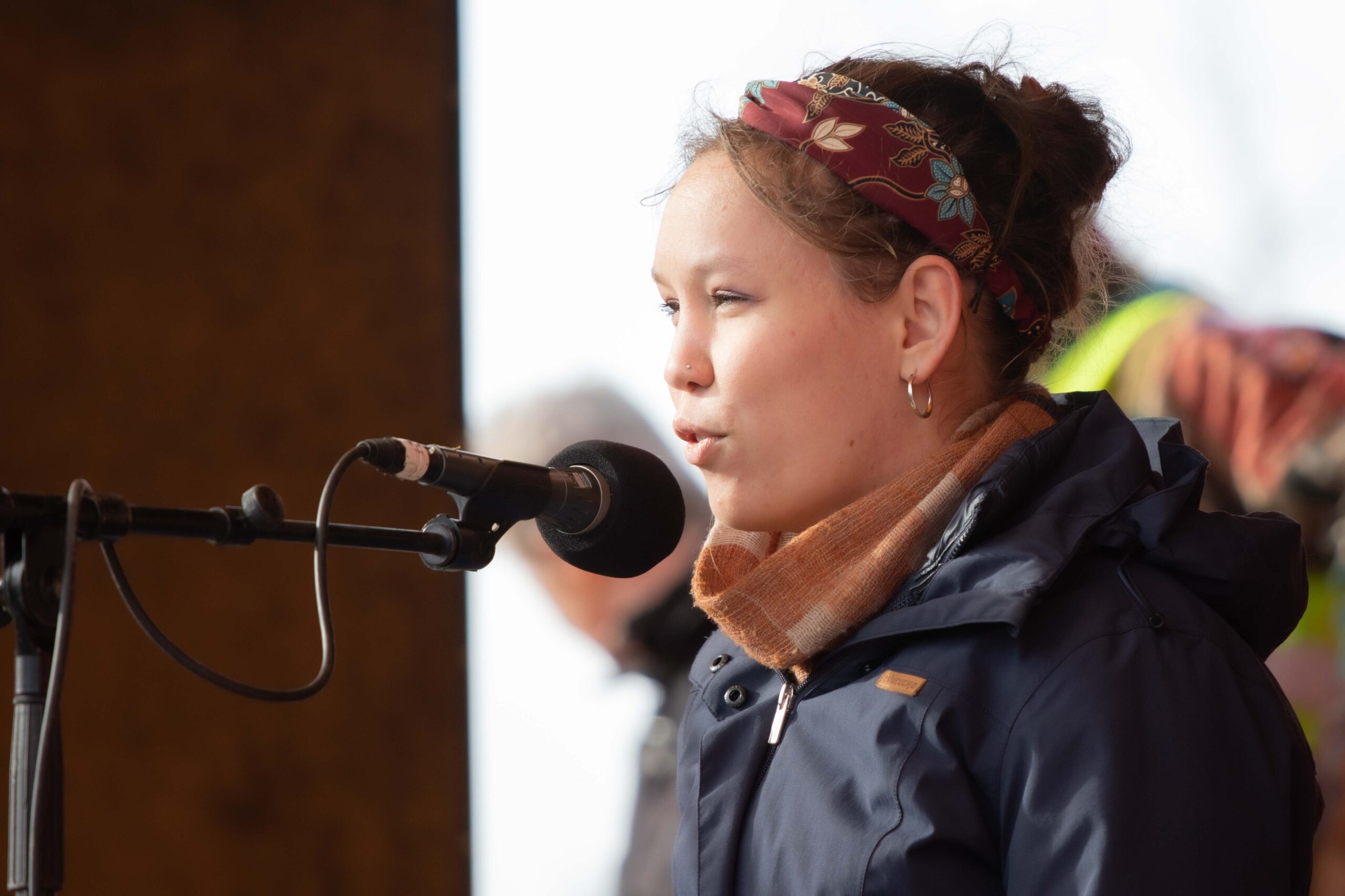 Foto door Jens van Hecke; ik zit rechts in beeld en praat naar links in een microfoon. Ik ben een indo vrouw met opgestoken donker haar met een batik haarband erin. Ik draag een oranje sjaal en donkerblauwe jas. 