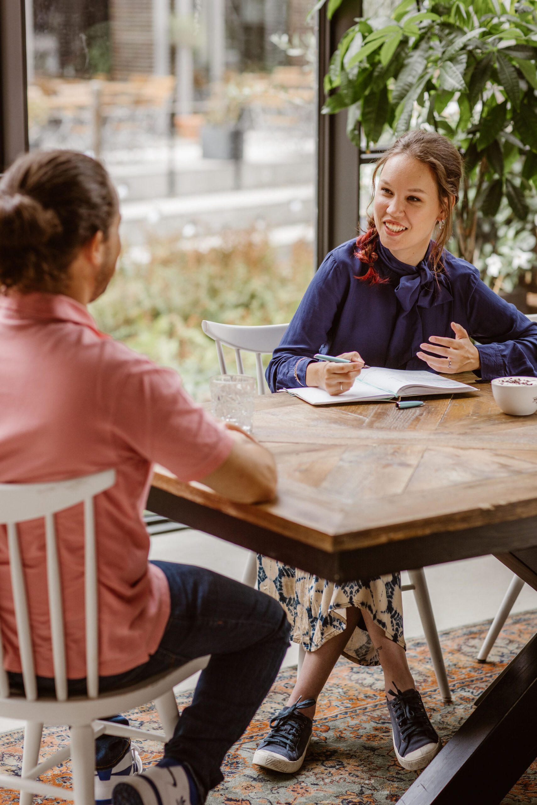 Foto door Marijn de Wijs; Ik zit rechts in beeld achter een houten tafel en praat naar een man links met zijn rug naar de tafel. De man heeft donker haar in een knot en draagt een spijkerbroek met roze polo, hij is wazig in beeld en kijkt naar mij. Ik ben een indo vrouw met lang haar dat in een soort vlecht over mijn schouder valt, ik draag een donkerblauwe blouse met strik, een pen in mijn hand en een notitieboek voor mij op tafel. 