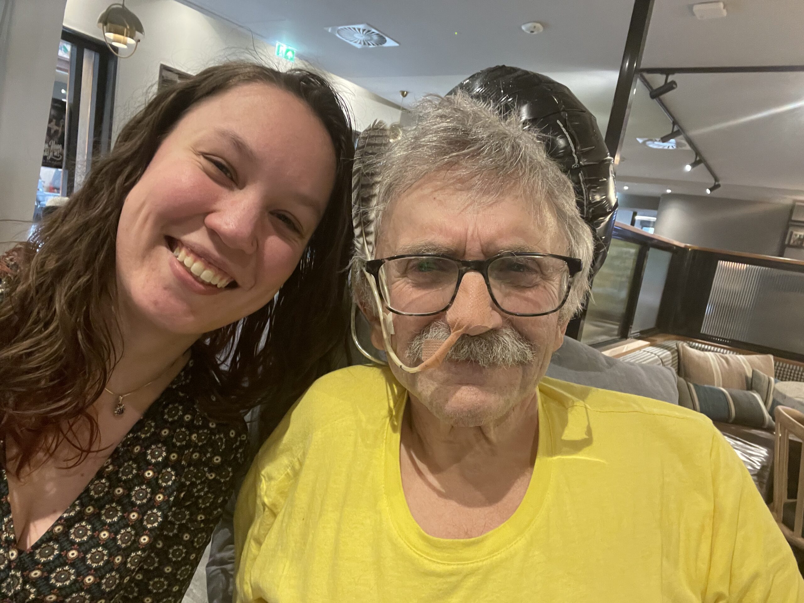 Selfie van mijzelf met Jan Troost, we lachen naar de camera. Ik ben een indo vrouw met lang roodbruin haar, Jan is een witte man met warrig grijs haar en een grijze snor, hij draagt een bril met zwart montuur waarachter een grijs-wit-zwarte veer gestoken is, in zijn neus zit een sonde.