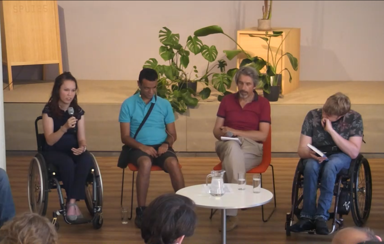 screenshot uit "de sluier van onwetendheid", panelgesprek. Jiska (indo vrouw in rolstoel), Ihab (blinde marrokaanse man op stoel), Machiel (witte man op stoel), Maurits (witte man in rolstoel) zitten om een kleine tafel met glazen water. Voor in beeld een paar stukjes achterhoofd van toeschouwers.