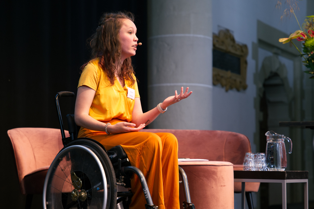 Foto door Tiva Pam, van mij; ik praat naar links in beeld, zit in een rolstoel en gebaar met mijn handen. Ik ben een indo vrouw met half opgestoken donker haar en draag een goudgele glimmende blouse en oranje soepelvallende broek.