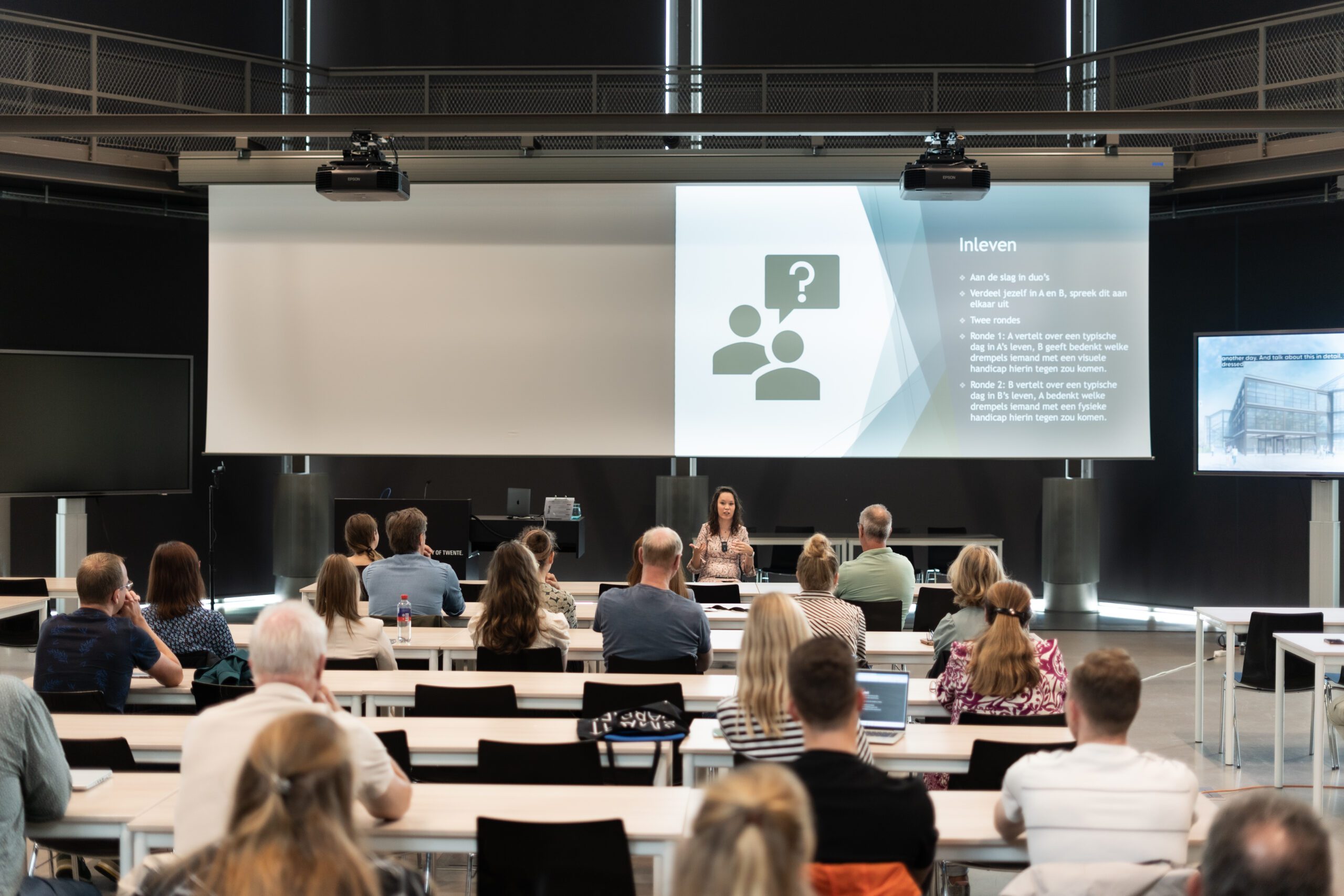 Foto door Bo ten Broeke//Annabel Jeuring. Zaal van achter gefotografeerd, er zitten mensen in de zaal. Vooraan tussen de mensen door zie je mij; een indo vrouw met halflang donker haar in een roze jurk met print in een rolstoel. Achter/boven mij een scherm met daarop mijn powerpoint.