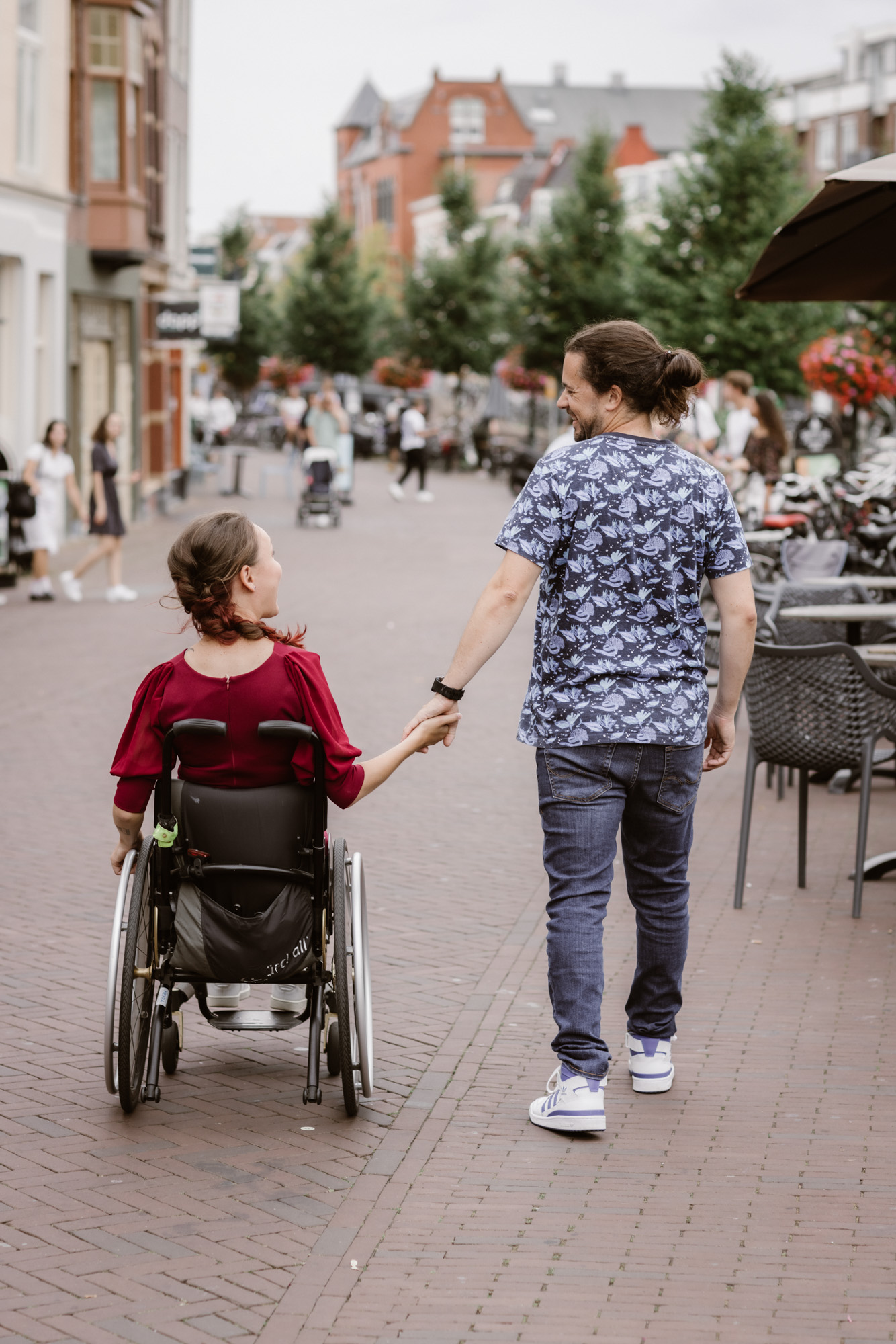 Foto door Marijn de Wijs; straatbeeld met twee mensen van de rug gefotografeerd: links ikzelf, ik zit in een donkere rolstoel, draag een bordeauxrode jumpsuit en witte sneakers. Mijn lange donkere haar zit in een twist, ik ben een indo vrouw. Rechts, hand in hand mijn partner. Hij is een witte man met lang donker krullend haar in een knot en een baardje. Hij draagt een shirt met blauwe print, een blauwe spijkerbroek en witte sneakers met blauw detail. We kijken lachend naar elkaar.