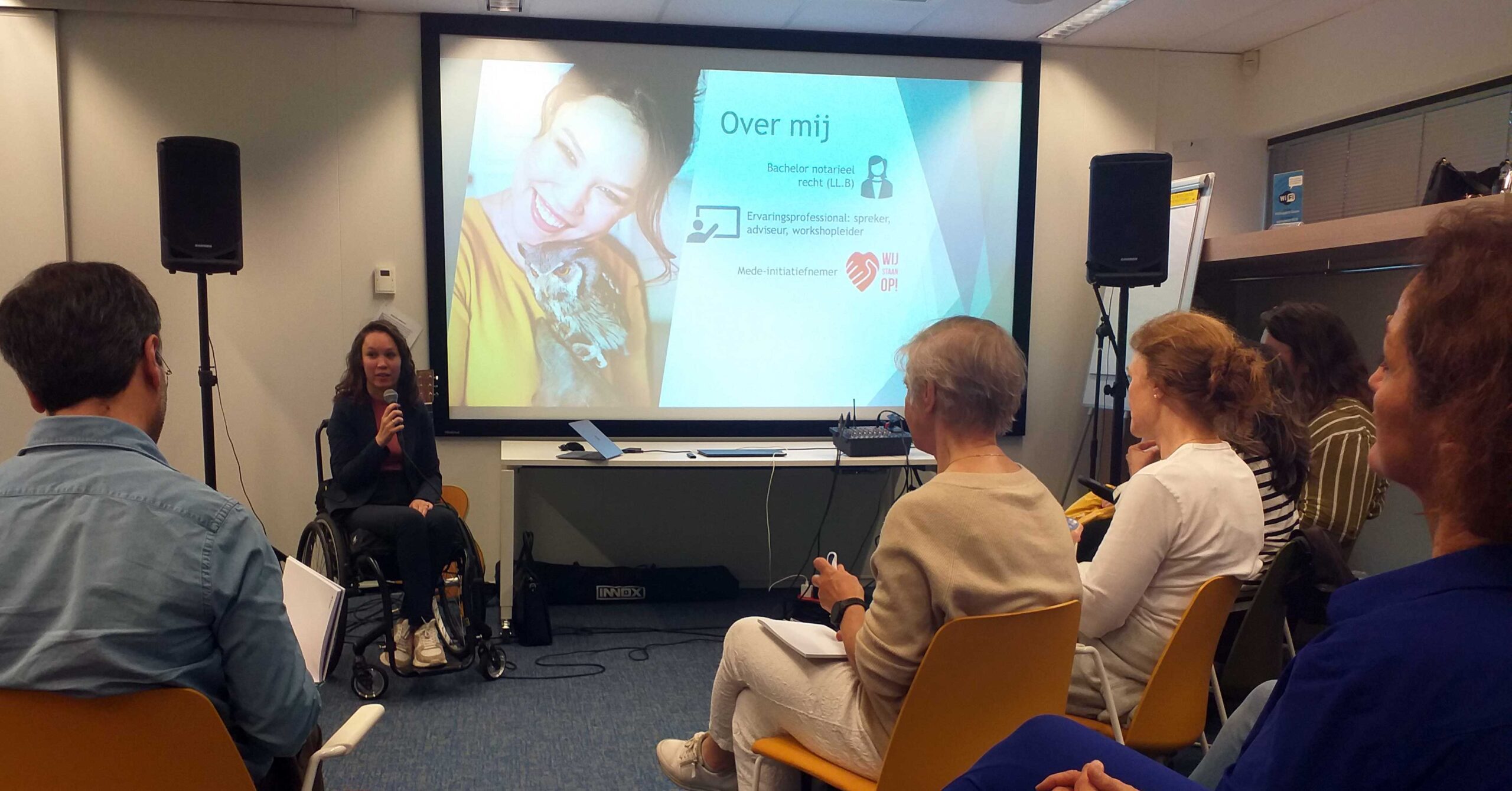 Foto genomen van achterin een zaal mensen. Vooraan hangt een groot scherm met daarop mijn powerpoint-presentatie. Ik sta links schuin voor het scherm. Ik ben een indo vrouw met donker golvend haar in een zwarte handbewogen rolstoel. Ik draag een donkerblauw soepelvallend pak en een roze col. In mijn hand heb ik een microfoon en ik richt mij tot de zaal.