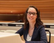 Foto door Jens Van Hecke: ik zit in mijn rolstoel achter de crèmewitte desk van een VN-zaal. Ik ben een indo vrouw met donker golvend haar en een bruin montuur bril op. Ik draag een witte blouse met glinsterend zilveren bies en een donkerblauw colbert. Ik kijk schuin lachend naar de camera, links van mij is een microfoon te zien.