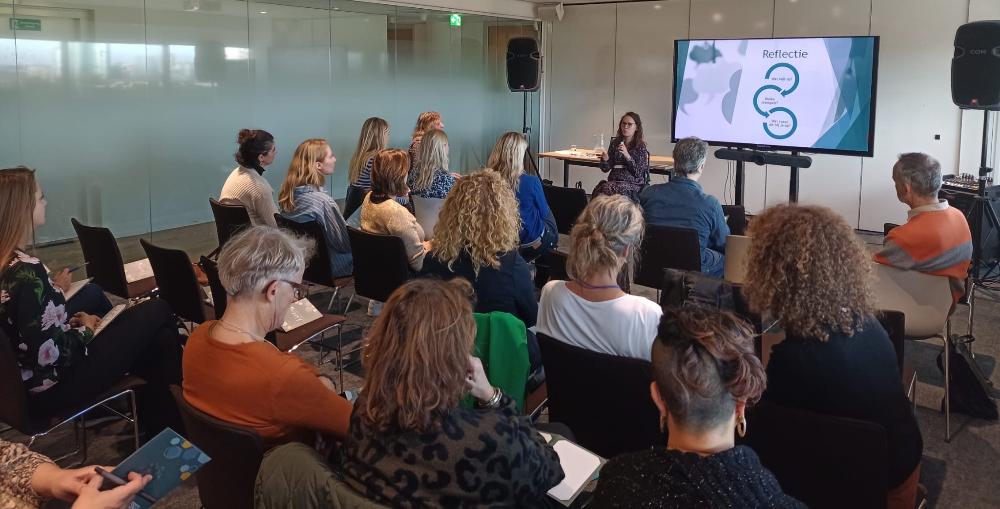 Jiska Vilans Foto door Jeroen Havers, schuin achterin de zaal genomen. Je ziet de ruggen en zijkanten van publiek, vooraan de zaal sta ik. Ik ben een indo vrouw met donker golvend haar en een donker montuur bril, ik zit in een zwarte rolstoel en houd een microfoon vast.