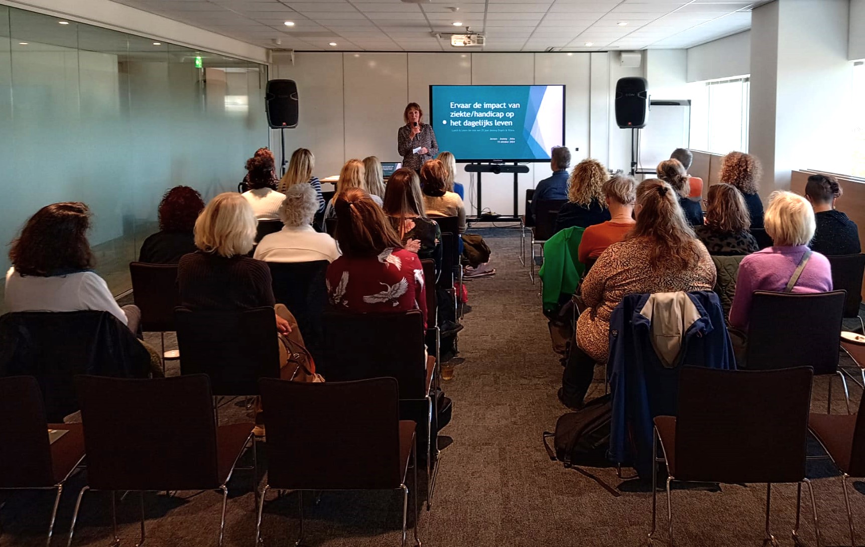 Jeanny Foto door Jeroen Havers, genomen achterin de zaal. In beeld zijn de ruggen van het publiek, vooraan staat Jeanny Engels voor de presentatie. Jeanny is een witte vrouw met bruin haar en draagt een blouse met motief en houd de microfoon in haar rechter hand. In haar linker hand heeft ze een klein briefje.