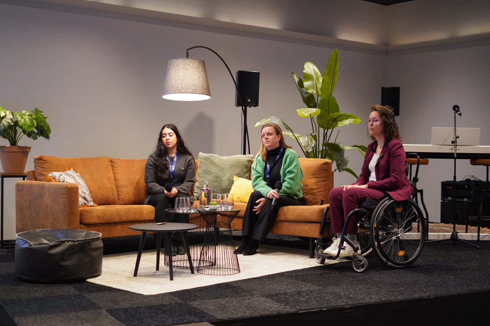 Foto door Iris Molenaar: podium met daarop een bruinkleurige bank. Rechts naast de bank zit ik in een zwarte rolstoel. Ik ben een indo vrouw met bruin golvend haar en een dun bruin halfrond montuur bril. Ik draag witte sneakers en een wit shirt met een bordeauxrood pak en een speld van een uil op mijn revers. In het midden zit Romke Biagioni, een witte vrouw met blond haar, zwarte broek en shirt, en een groene wollen trui. Links zit Nour Al Kuhaili, een jonge vrouw van kleur met donker lang krullend haar, ze draagt een zwarte broek en een grijs gebreid vest.