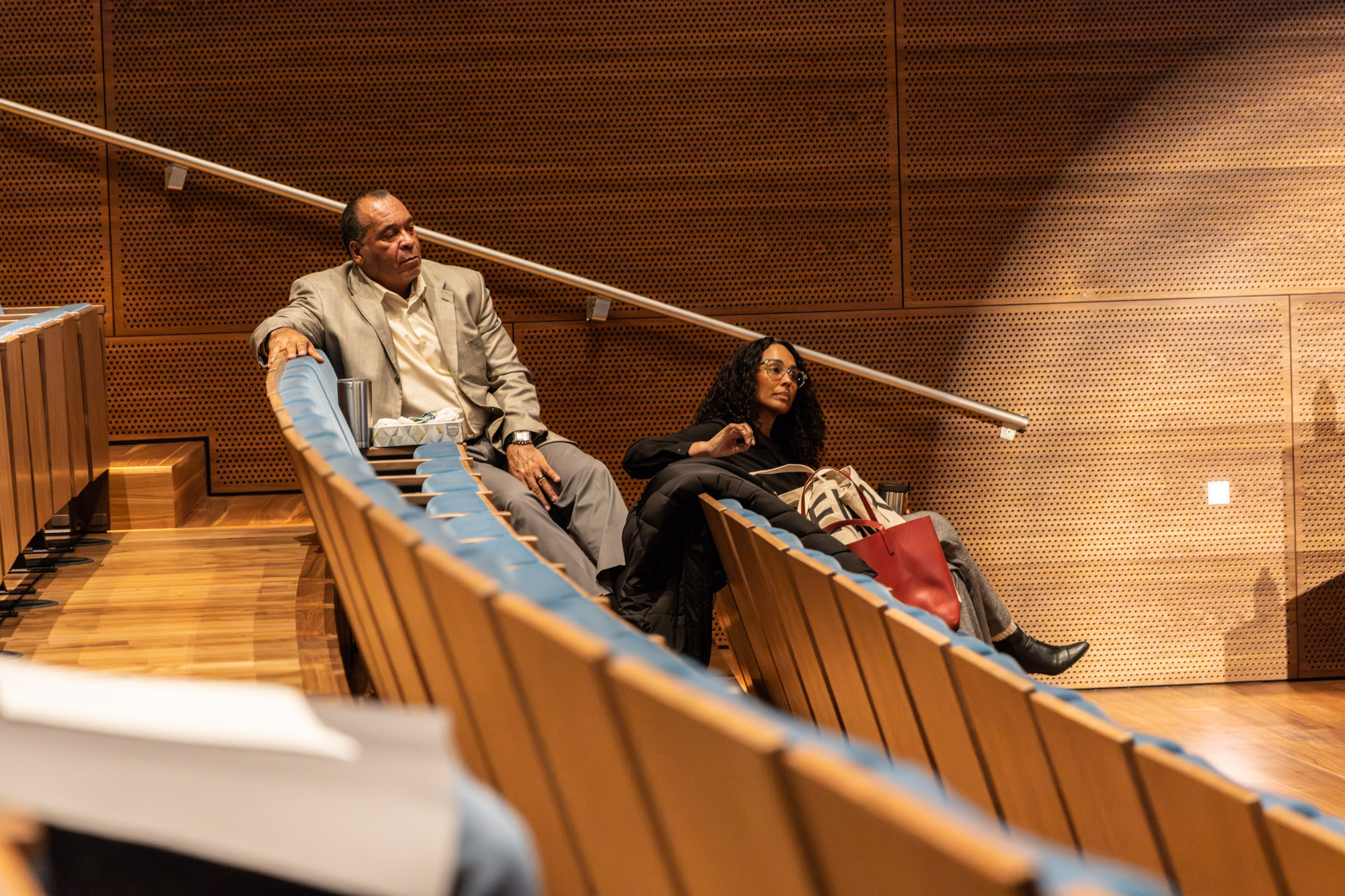 photos by Jens Van Hecke; insta @Jensvh_photos; jensvanhecke@hotmail.com Foto door Jens Van Hecke: deel van de theaterzaal met achterin twee toehoorders. Larry, een donkere man die een wit overhemd en grijs colbert draagt. En Leeza, een donkere vrouw met donkere krullen en een transparant groot monuur bril. Ze kijken naar rechts buiten beeld, waar de sprekers staan.