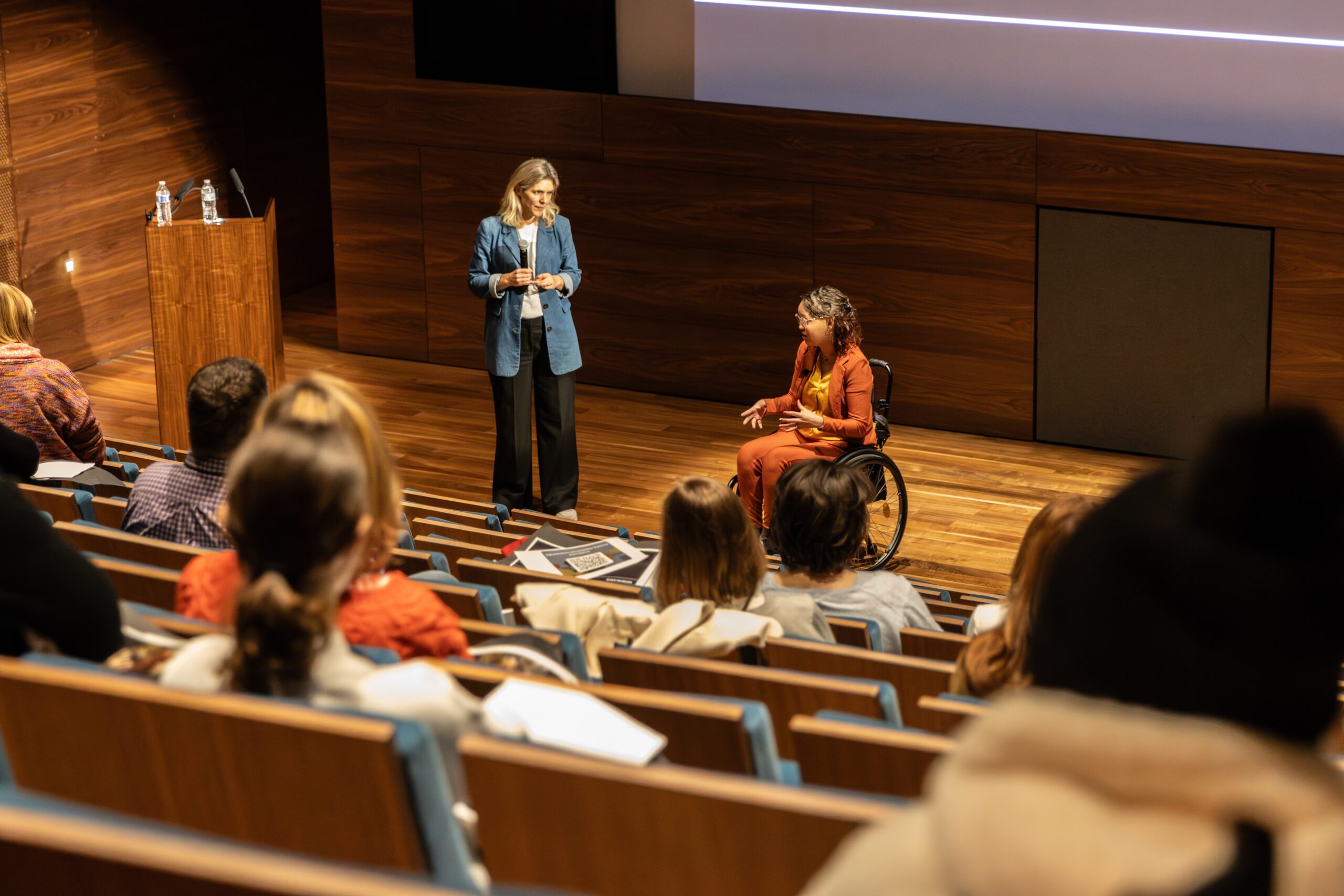Foto door Jens Van Hecke: donkere zaal met op de voorgrond een theateropstelling van stoelen waar deels mensen in zitten. Beneden op het podium staan Cathelijne en Jiska. Cathelijne is een witte vrouw met blond haar, ze draagt een donkere broek, een wit T-shirt en een denim-blauw lang colbert, ze kijkt naar Jiska. Jiska is een indo vrouw met donker golvend schouderlengte haar in een zwarte rolstoel. Ze draagt een okergele top met een oranje pak en een transparante bril.