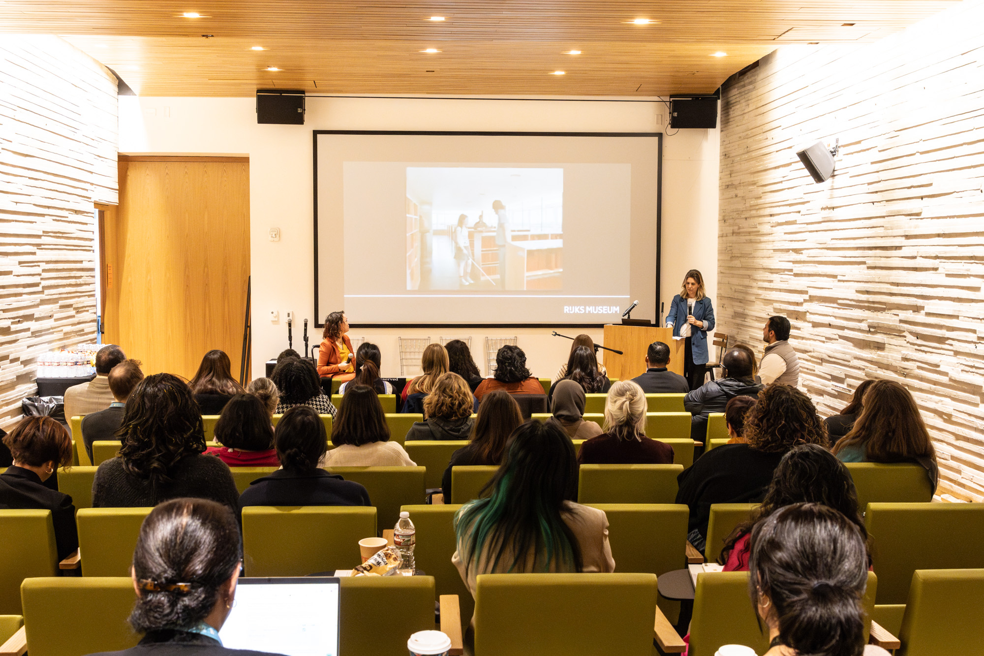 photos by Jens Van Hecke; insta @Jensvh_photos; jensvanhecke@hotmail.com Foto door Jens Van Hecke: symposiumzaal van achterin gefotografeerd. Op de voorgrond mensen in stoelen, op de achtergrond van de foto een groot scherm met een PowerPointpresentatie, links in beeld ben ik te zien. Een indo vrouw met donker golvend haar in een zwarte rolstoel met een oranje pak. Rechts in beeld Cathelijne, een witte vrouw met blond haar, een donkere broek, wit T-shirt en een denimblauw lang colbert.