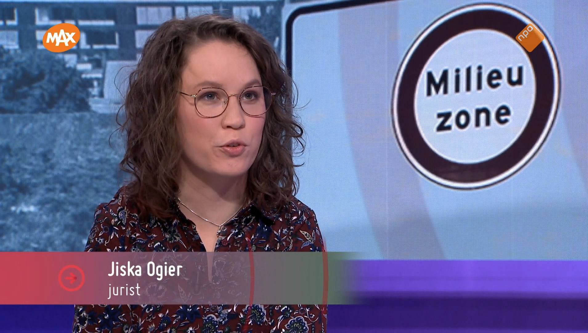 Afbeelding uit de uitzending van Max Meldpunt. Ik zit in de studio, over mijn borst een balkje met mijn naam en "jurist", op de achtergrond een bord van een milieuzone. Ik ben een indo vrouw met golvend donker haar, ik draag een blousejurk met patroon en een dun bruin montuur bril, ik praat naar rechts buiten beeld.