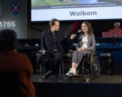 Foto door Erik Kottier: Ik zit in mijn rolstoel op een podum, naast mij zit Stan Verhaag op een stoel en houdt mij een microfoon voor. Op de donkere voorgrond zitten mensen met rug naar de camera, waaronder Illya links. Ik ben een indo vrouw met donker golvend haar en een dun bruin montuur bril, ik zit in een zwarte rolstoel en draag een donkerblauw pak met wit bloemenpatroon, een witte soepele blouse en witte sneakers. Stan is een witte man met kort bruin haar, hij draagt een donker geruit pak en donkere blouse.