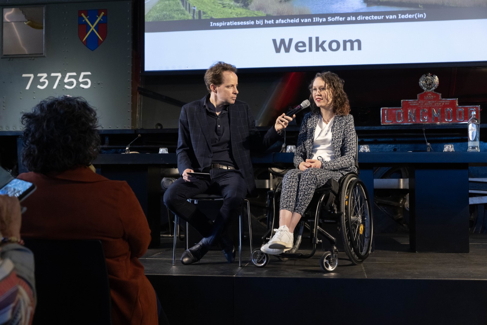 Foto door Erik Kottier: Ik zit in mijn rolstoel op een podum, naast mij zit Stan Verhaag op een stoel en houdt mij een microfoon voor. Op de donkere voorgrond zitten mensen met rug naar de camera, waaronder Illya links. Ik ben een indo vrouw met donker golvend haar en een dun bruin montuur bril, ik zit in een zwarte rolstoel en draag een donkerblauw pak met wit bloemenpatroon, een witte soepele blouse en witte sneakers. Stan is een witte man met kort bruin haar, hij draagt een donker geruit pak en donkere blouse.