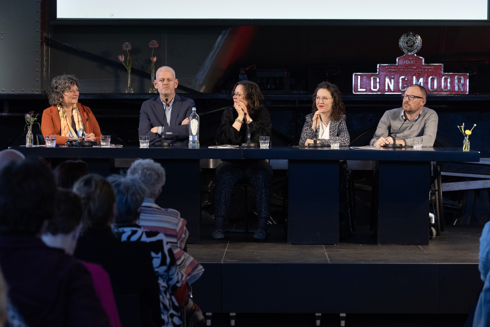 Foto door Erik Kottier: doorkijkje tussen de gasten naar het podium. Achter een desk op het podium zitten van links naar rechts: Stan Verhaag (dagvoorzitter), Illya Soffer (afzwaaiend directeur), Albert-Jan Kruiter, Tamara van Ark, ikzelf, Otwin van Dijk (allen panellid). 