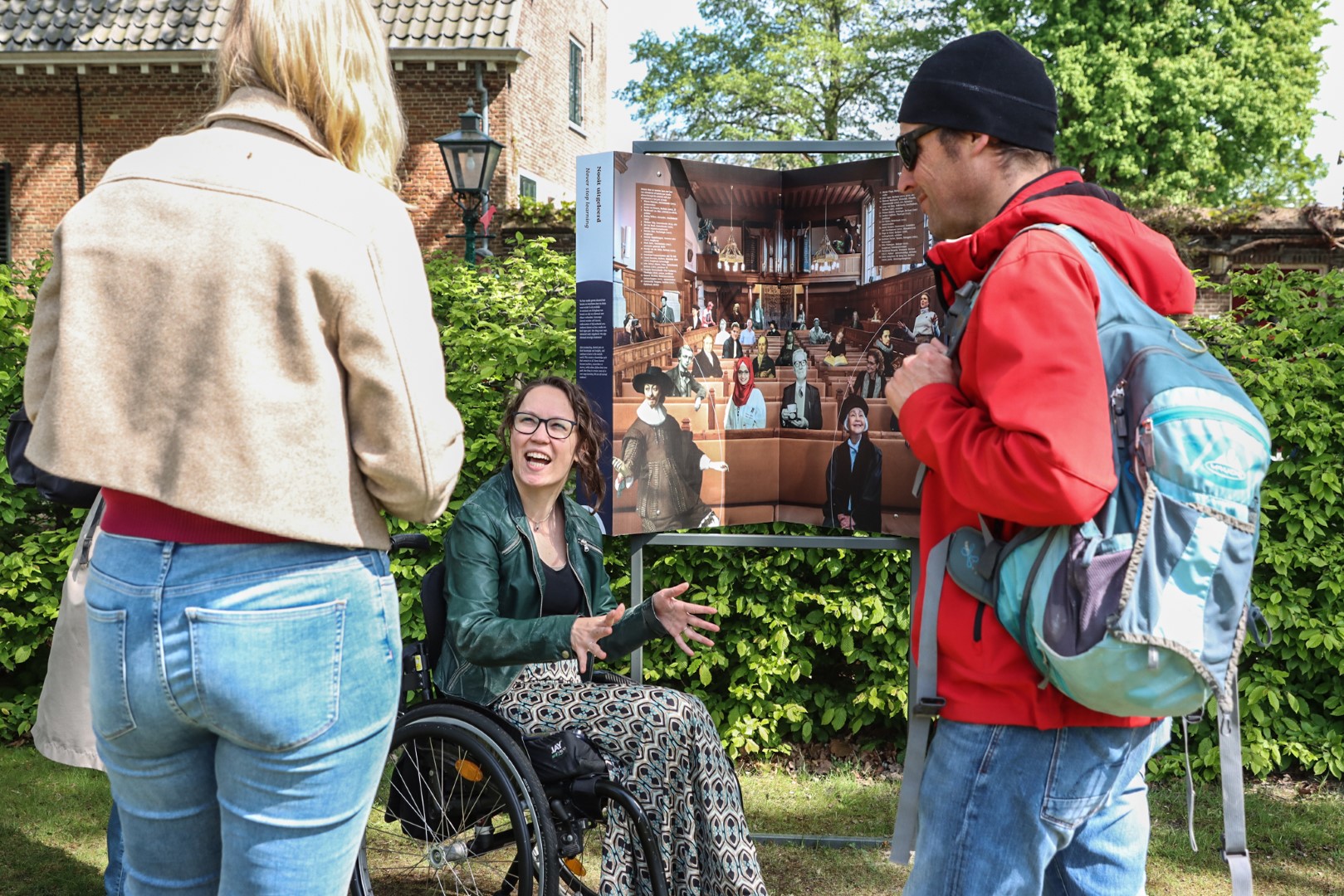 Foto door Jens Van Hecke: een aantal mensen staan bij de tentoonstelling buiten, voor de beeldredactie het Auditorium. Ik sta voor de plaat en praat met de kijkers. Ik ben een indo vrouw met donker golvend opgestoken haar, ik zit in een zwarte rolstoel en draag een gedesigneerde rok, zwart T-shirt en donkergroene leren jas. 